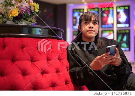 Asian Person Seated on Plush Couch Holding Smartphone Asian Person Seated on Plush Couch Holding Smartphone 141726975