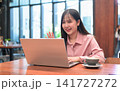Smiling Asian Woman Waving at Her Laptop Screen during a Video Call 141727272
