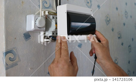 The process of installing an electrical panel on a wallpapered wall. Hands position the white plastic housing with a dark protective cover over the circuit breakers, holding a screwdriver nearby. 141728106