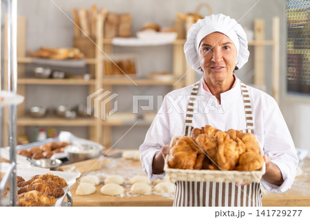 Mature smiling woman baker showing warm tasty croissants baked in small bakery Mature smiling woman baker showing warm tasty croissants baked in small bakery 141729277