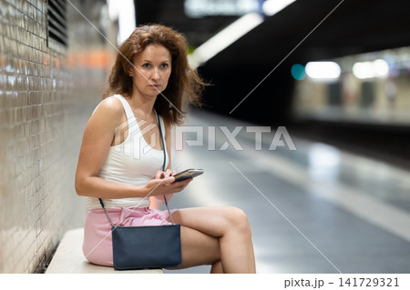 Woman looking for subway train schedule on smartphone while sitting on bench at underground metro station Woman looking for subway train schedule on smartphone while sitting on bench at underground metro station 141729321
