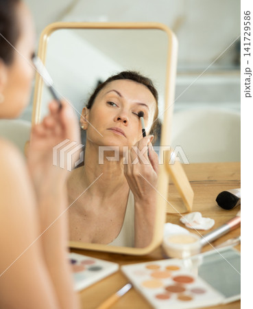 Middle-aged woman using brush during eye makeup sitting in front of the mirror Middle-aged woman using brush during eye makeup sitting in front of the mirror 141729586