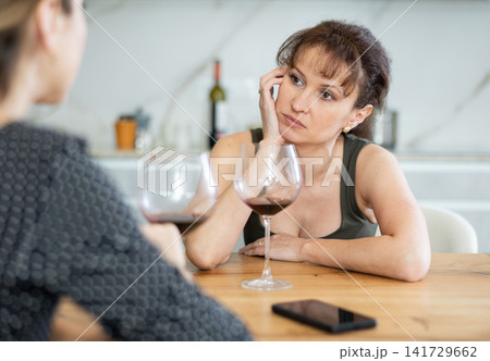 Middle-aged woman sitting at table with a glass of wine and talking to another at home Middle-aged woman sitting at table with a glass of wine and talking to another at home 141729662