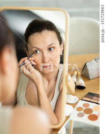 Middle-aged woman doing eye make-up looking in the mirror while seated at a table Middle-aged woman doing eye make-up looking in the mirror while seated at a table 141729663