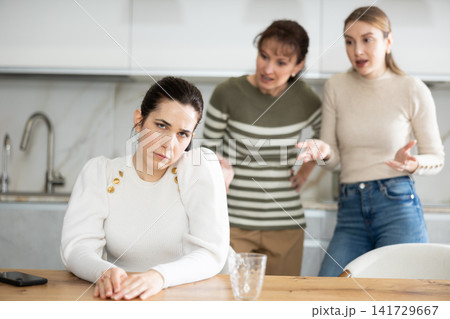 Upset middle-aged woman sitting at the kitchen table with her back to women quarreling to her 141729667