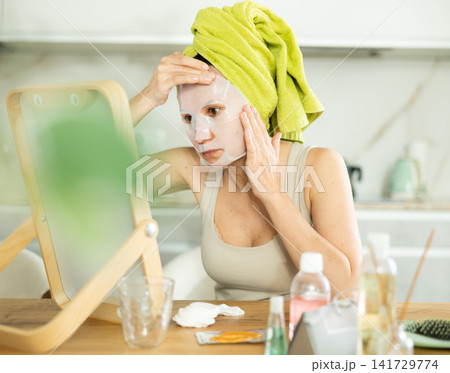 Middle-aged woman applying tissue mask on her facial skin sitting in front of the mirror Middle-aged woman applying tissue mask on her facial skin sitting in front of the mirror 141729774