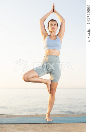 Sedulous woman doing warrior one leg pose of yoga on mat on beach at sunrise 141729866