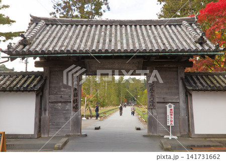 A traditional wooden gate stands at the entrance of a serene pathway, flanked by autumn foliage and visitors strolling under clear skies. 141731662