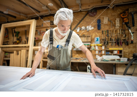Professional man restorer carefully examines painted antique interior door on workbench in workshop. Professional man restorer carefully examines painted antique interior door on workbench in workshop. 141732815