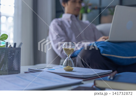 Man working on laptop sitting on sofa at home with running hourglass on the table. Concept for time management and extra over time working. 141732872