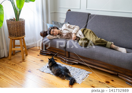 Lazy happy woman peacefully relaxing on couch and observing domestic cat resting on rug in room. 141732989