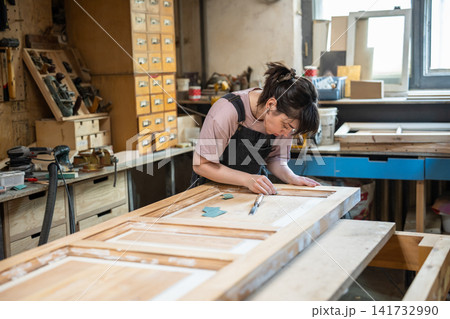Interested concentrated woman crafter refurbishing vintage wooden door in restoration workshop. Interested concentrated woman crafter refurbishing vintage wooden door in restoration workshop. 141732990