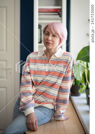 Relaxed positive stylish mature woman posing at camera while sitting on table beside window at home. 141733000