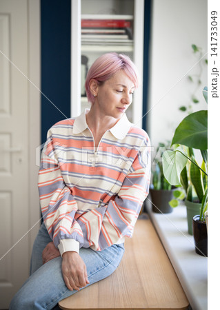 Smiling calm mature woman observing indoor windowsill garden while sitting on table beside window. Smiling calm mature woman observing indoor windowsill garden while sitting on table beside window. 141733049