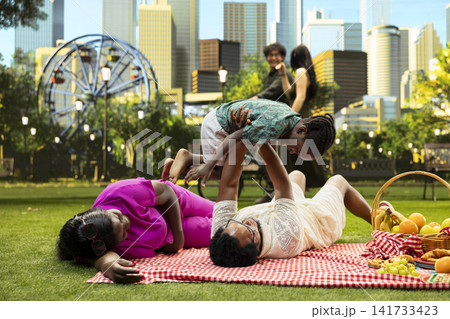 Father lifting child in airplane play during family picnic in park, parents laying on picnic blanket and playing with young boy beside basket of fruits and pastries. 141733423