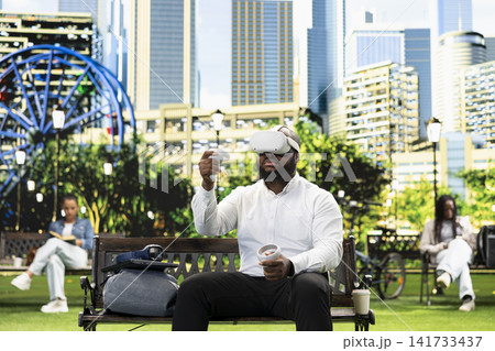 Businessman with virtual reality headset and motion controllers on park bench, with pedestrians in green background. VR glasses immersive simulation with tech. Businessman with virtual reality headset and motion controllers on park bench, with pedestrians in green background. VR glasses immersive simulation with tech. 141733437
