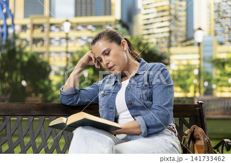 Casually dressed woman leans on one hand and reads open book on wooden bench in urban park, portraying concentration, calm and mindful leisure. Book lover recreation. Casually dressed woman leans on one hand and reads open book on wooden bench in urban park, portraying concentration, calm and mindful leisure. Book lover recreation. 141733462