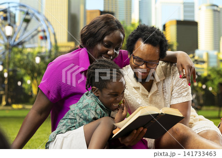 African parents and child reading book together on picnic blanket, sharing calm learning and storytelling moment beside picnic arrangements in sunny city park. Weekend fun. African parents and child reading book together on picnic blanket, sharing calm learning and storytelling moment beside picnic arrangements in sunny city park. Weekend fun. 141733463