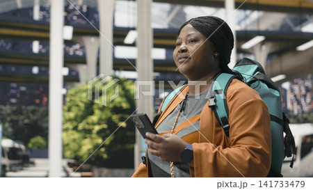 Close up of smiling woman in bus terminal using smartphone travel app while walking with packed backpack. Joyous passenger reads journey information on mobile phone screen Close up of smiling woman in bus terminal using smartphone travel app while walking with packed backpack. Joyous passenger reads journey information on mobile phone screen 141733479