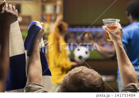 Close up of football supporters raising beer cup and scarf while watching soccer game on television. Group of friends with alcoholic beverage and team colors fan props in hand Close up of football supporters raising beer cup and scarf while watching soccer game on television. Group of friends with alcoholic beverage and team colors fan props in hand 141733484