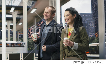 Smiling business travelers walk together through bus terminal with coffee cups in hand. Joyous businesspeople enjoying takeaway drinks in shuttle station, discussing travel itinerary 141733525
