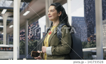 Smiling passenger holding mobile device while continuing through bus terminal. Jolly solo traveler checks digital route information on smartphone while walking in shuttle station 141733528