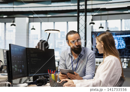 Senior engineer mentoring junior developer at workstation in office, reviewing AI system metrics. Tech company experienced manager guiding intern through code reviewing using machine learning 141733556