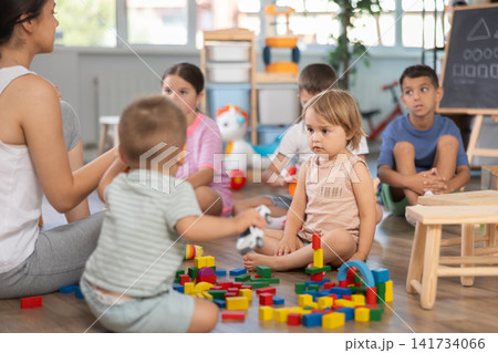Caring young woman teacher playing educational games with group of children sitting on floor at daycare Caring young woman teacher playing educational games with group of children sitting on floor at daycare 141734066