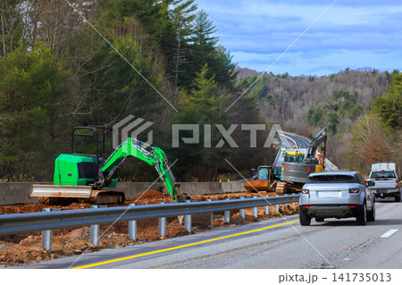 Excavators work on side of highway while cars drive nearby in wooded area Excavators work on side of highway while cars drive nearby in wooded area 141735013