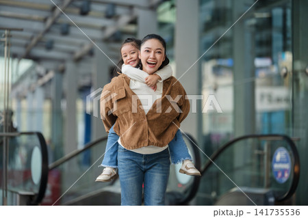 happy mother giving daughter piggyback ride near escalator in shopping mall or department store happy mother giving daughter piggyback ride near escalator in shopping mall or department store 141735536