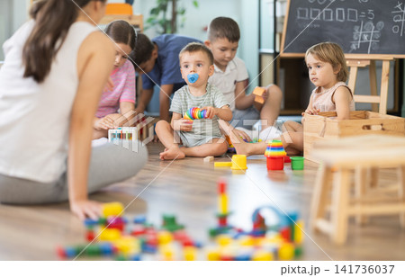 Young experienced young female teacher supporting to children sitting on floor and spending playing time at classes for kids Young experienced young female teacher supporting to children sitting on floor and spending playing time at classes for kids 141736037