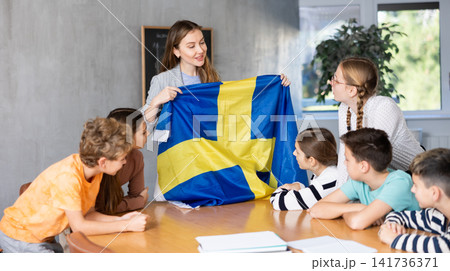 Group of curious preteen learners and young female teacher with flag of Sweden in classroom of academy 141736371