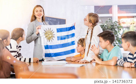 Cheerful young female teacher showing flag of Uruguay to schoolchildren preteens during history lesson in audience Cheerful young female teacher showing flag of Uruguay to schoolchildren preteens during history lesson in audience 141736372