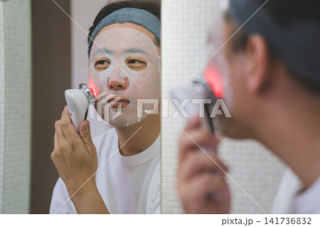 鏡の前でシートマスクをつけながらLED美顔器で頬を整える40代男性のスキンケア 141736832