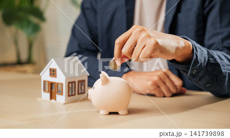 A businessman hand places a coin into a piggy bank beside a house model, symbolizing property savings, financial planning, and investment strategies for home buying 141739898