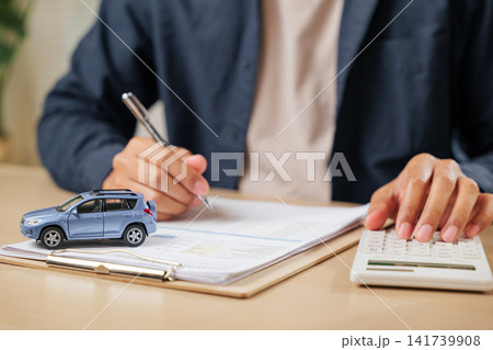 A man signs car insurance documents, calculating costs with a loan calculator. Writing his signature on a contract, ensuring smooth financial transactions A man signs car insurance documents, calculating costs with a loan calculator. Writing his signature on a contract, ensuring smooth financial transactions 141739908