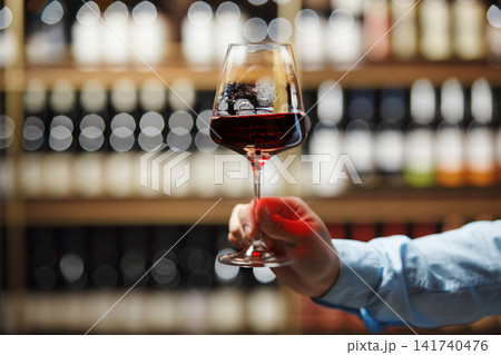 Close-Up of Sommeliers Hand Holding Glass of Red Wine Against Shelf of Assorted Bottles Close-Up of Sommeliers Hand Holding Glass of Red Wine Against Shelf of Assorted Bottles 141740476