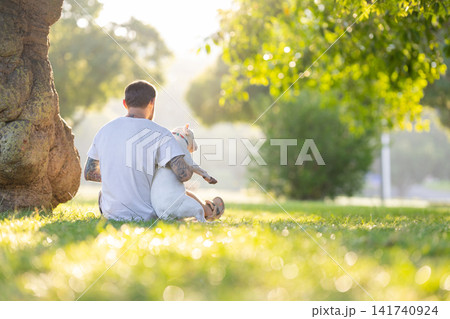 Old bull terrier sitting on the lap of a tattooed male owner in a green park, sunlight filtering through trees creating a serene outdoor atmosphere Old bull terrier sitting on the lap of a tattooed male owner in a green park, sunlight filtering through trees creating a serene outdoor atmosphere 141740924