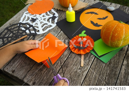 Little girl carefully cutting colored paper while making handmade Halloween decorations on a wooden table Little girl carefully cutting colored paper while making handmade Halloween decorations on a wooden table 141741583