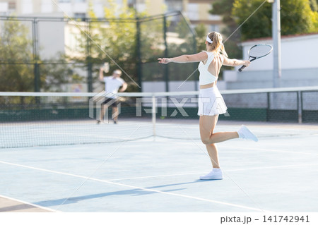 Woman plays tennis on a court while a man stands in the background at a sunny day in a sports facility 141742941