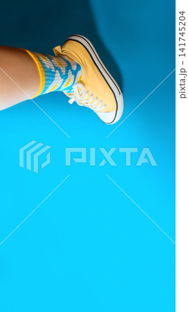 Leg wearing a yellow sneaker and colorful patterned socks against a vibrant bright blue background, modern fashion and youth lifestyle concept Leg wearing a yellow sneaker and colorful patterned socks against a vibrant bright blue background, modern fashion and youth lifestyle concept 141745204
