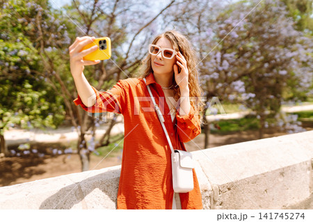 Young woman enjoys sunny day by taking selfie in vibrant park filled with blooming flowers. Blogging 141745274