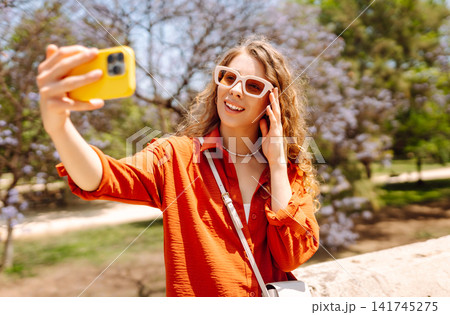 Young woman enjoys sunny day by taking selfie in vibrant park filled with blooming flowers. Blogging Young woman enjoys sunny day by taking selfie in vibrant park filled with blooming flowers. Blogging 141745275