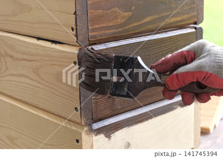 Hand in glove painting wooden box with brown oil stain brush. Worker applying protective varnish on pine timber planks surface, DIY woodwork. Wood staining process. Backyard Woodworking, Maintenance. Hand in glove painting wooden box with brown oil stain brush. Worker applying protective varnish on pine timber planks surface, DIY woodwork. Wood staining process. Backyard Woodworking, Maintenance. 141745394