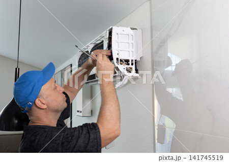 Technician man repairing air conditioning unit on wall. Professional maintenance worker using screwdriver. Climate control system service at home. Daytime soft lighting side view shot. Technician man repairing air conditioning unit on wall. Professional maintenance worker using screwdriver. Climate control system service at home. Daytime soft lighting side view shot. 141745519