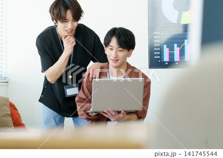 オフィスでノートパソコンを見ながら会話する若い日本人男性2人 141745544