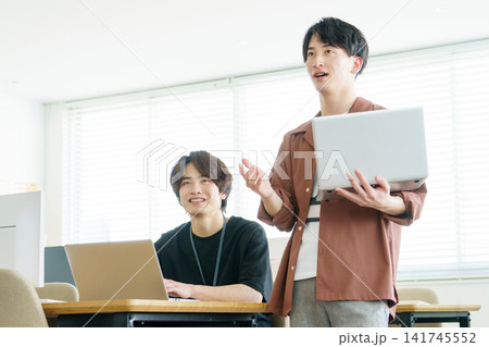オフィスでノートパソコンを見ながら会話する若い日本人男性2人 141745552