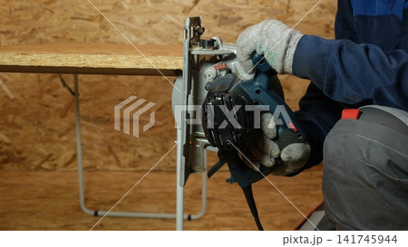 Builder saws a board with a circular saw cutting a wooden plank in a joinery. Carpenter uses portable circular saw tool to cut wood at wooden table in workshop. Worker sawing pine board. Real image Builder saws a board with a circular saw cutting a wooden plank in a joinery. Carpenter uses portable circular saw tool to cut wood at wooden table in workshop. Worker sawing pine board. Real image 141745944