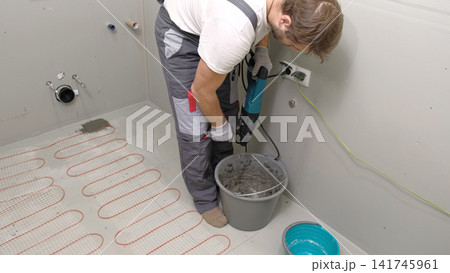 Construction worker preparing tile surface, mixing mortar substrate for flooring, underfloor heating mat. Workman combines adhesive in renovation setting. Operating power tool during bathroom upgrade Construction worker preparing tile surface, mixing mortar substrate for flooring, underfloor heating mat. Workman combines adhesive in renovation setting. Operating power tool during bathroom upgrade 141745961