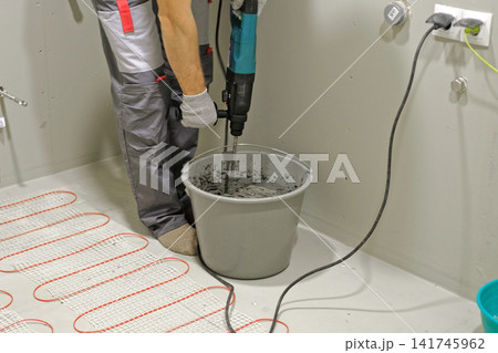 Construction worker preparing tile surface, mixing mortar substrate for flooring, underfloor heating mat. Workman combines adhesive in renovation setting. Operating power tool during bathroom upgrade Construction worker preparing tile surface, mixing mortar substrate for flooring, underfloor heating mat. Workman combines adhesive in renovation setting. Operating power tool during bathroom upgrade 141745962